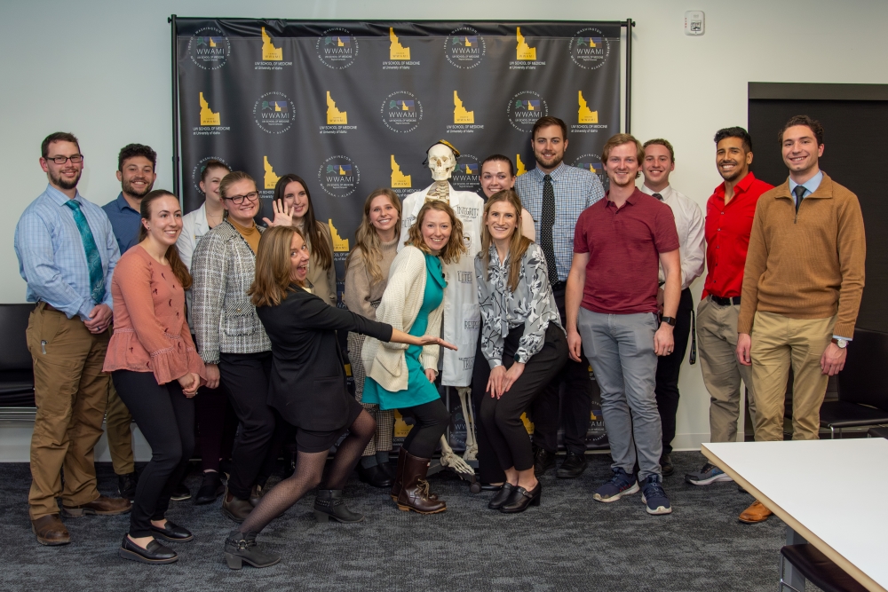 A group of Idaho WWAMI students pose in front of an black banner with a skeleton model. Dr. Fehrenkamp stands in front doing an exaggerated pose with her arms as though showcasing the students. 