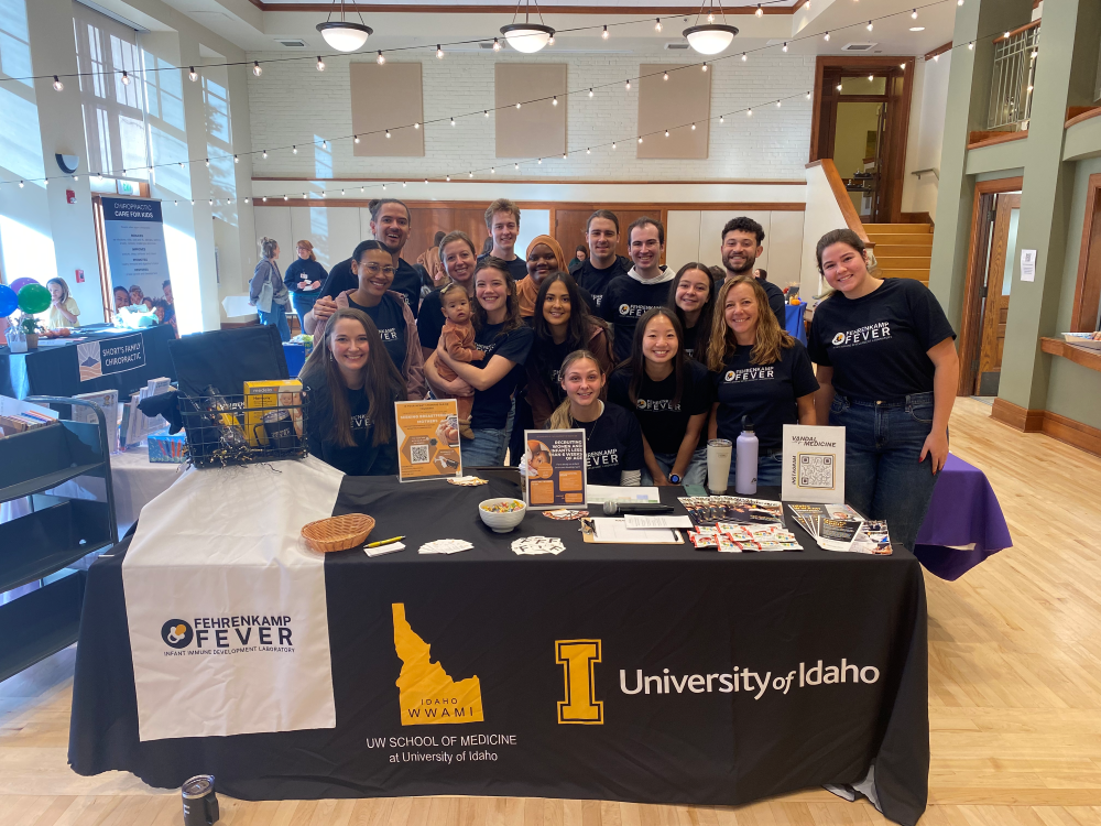 A group of people wearing "Fehrenkamp FEVER" T-shirts stand behind a table with Idaho WWAMI banners