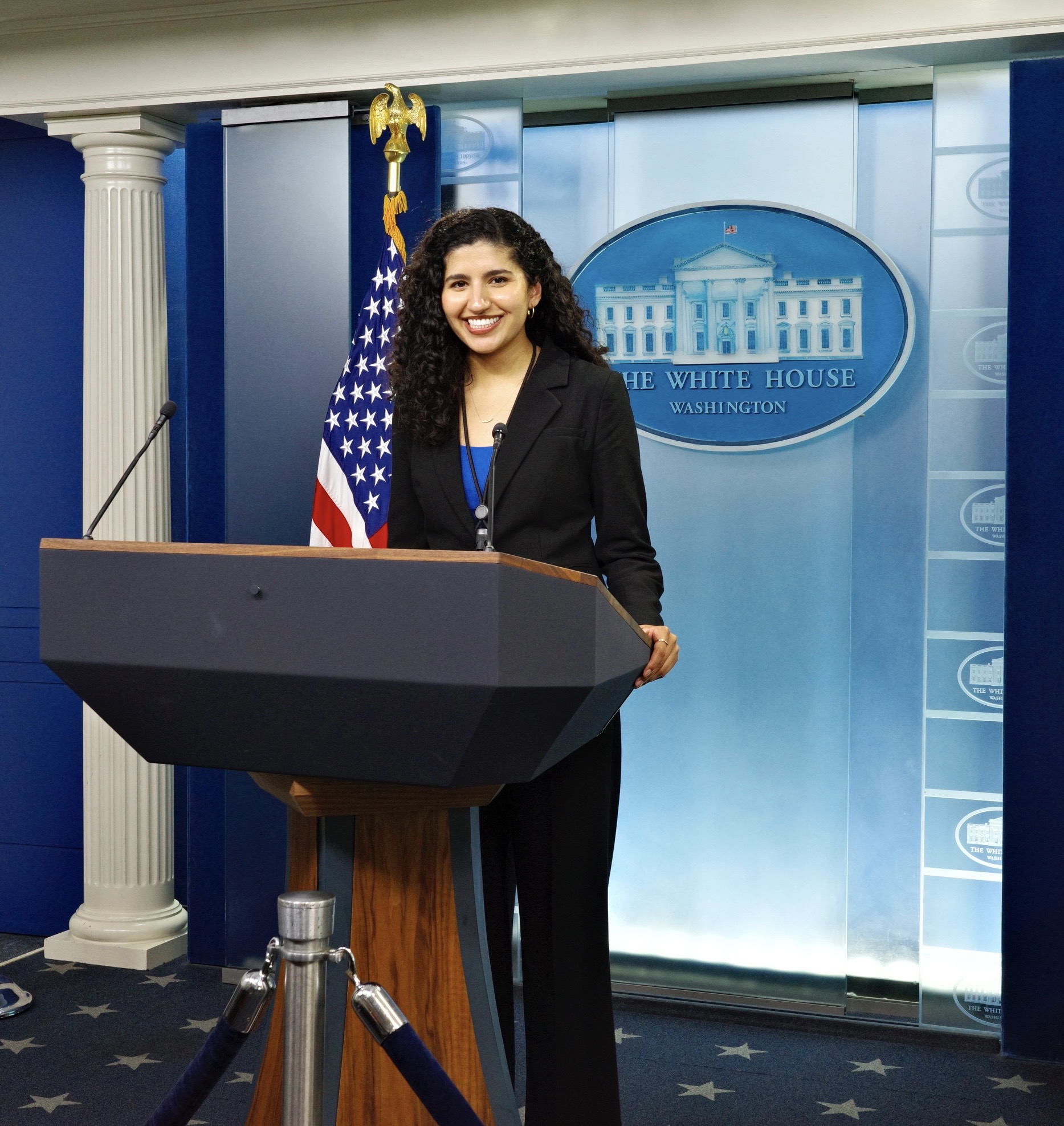 Briana Martin-Villa stands in front of a podium with the White House Insignia and an American Flag behind her.