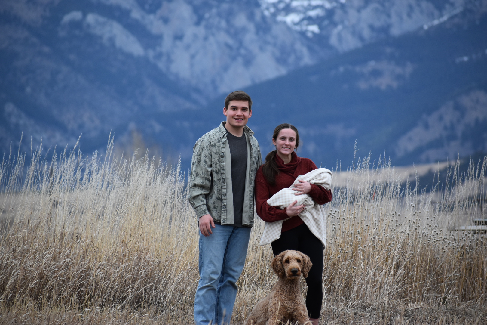 Ben and Olivia held pose in front of a Montana mountain range. Olivia is holding baby Winston and a dog poses at their feet. 
