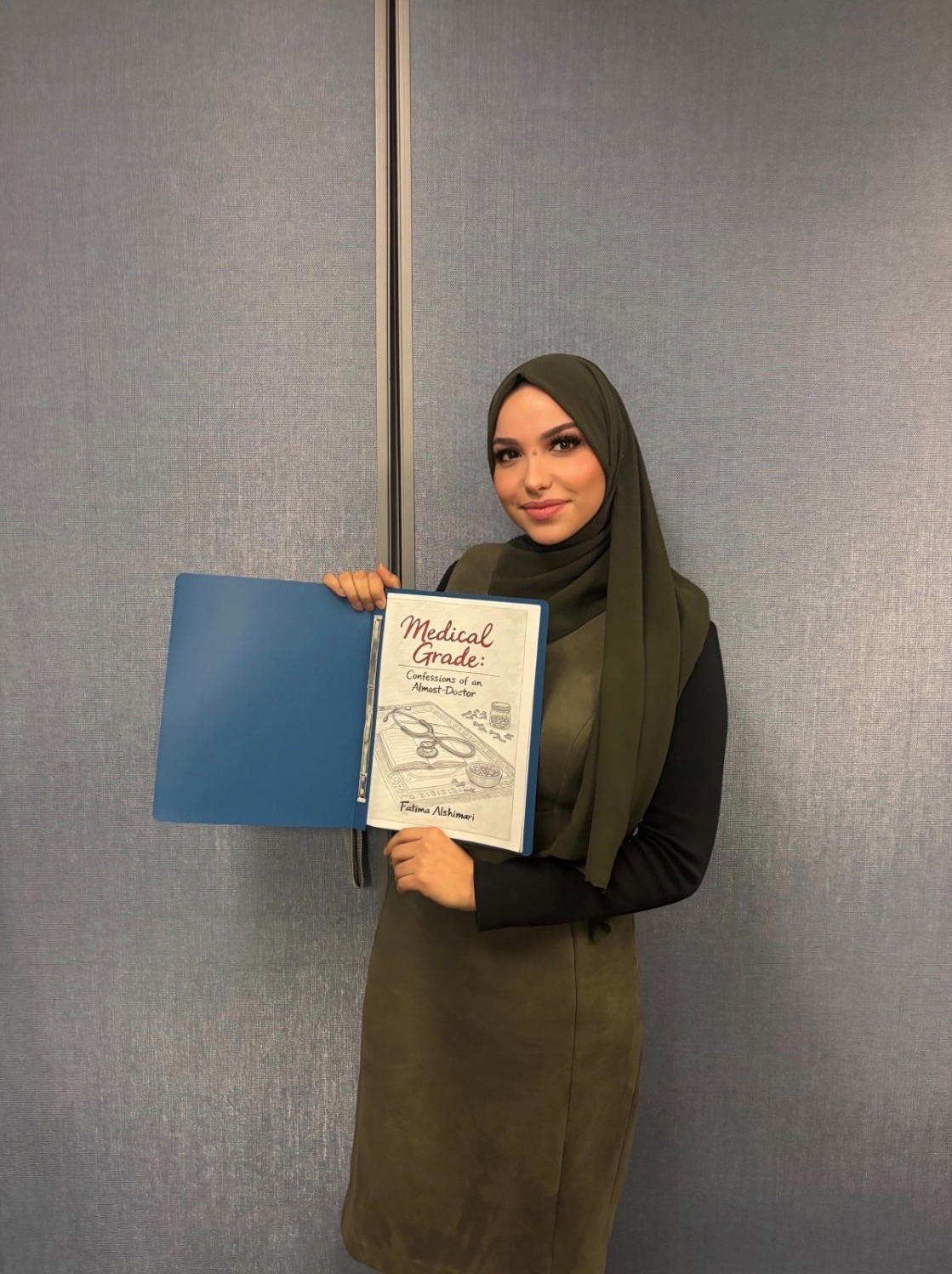 Fatima Al-Shimari poses with her project, a book with a blue cover, in front of a grey wall
