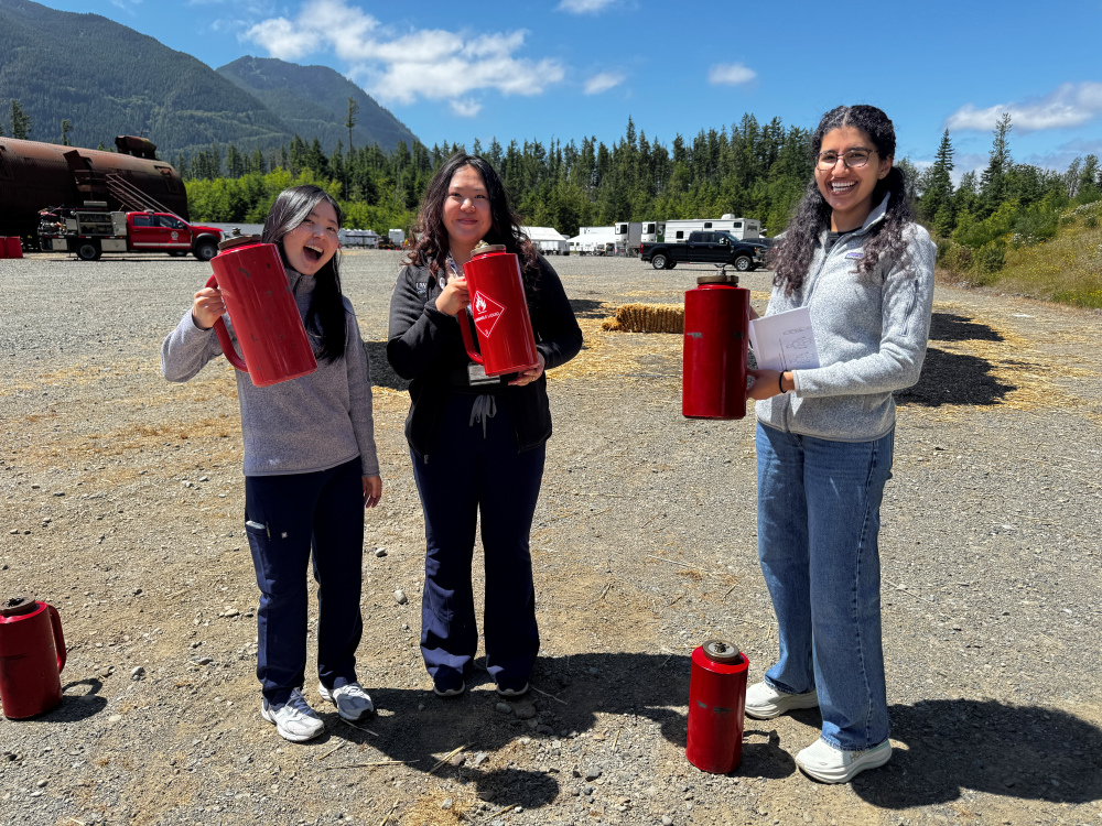 Three students stand in an open field holding fire cans